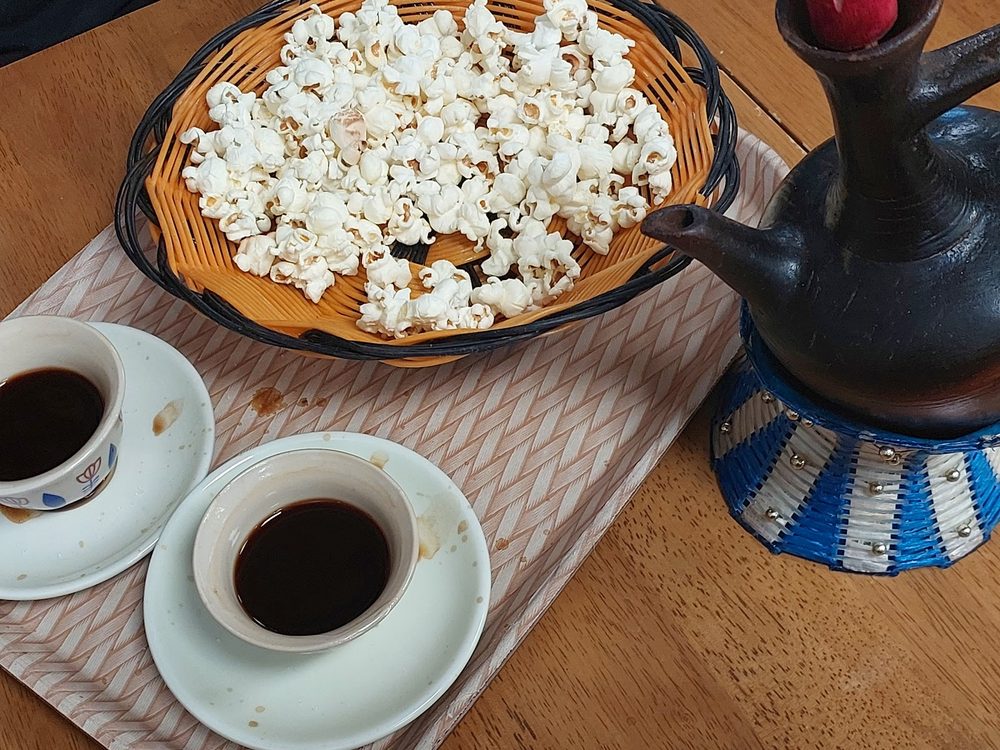 A bowl of popcorn beside a black clay jebena coffee pot and two white cups of black coffee on a woven mat.
